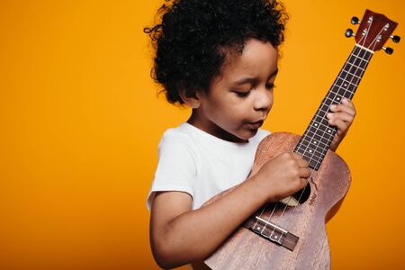 Dark-skinned, talented boy is fingering strings of a ukulele guitar, carefully listening to the sounds on an orange background.の写真素材