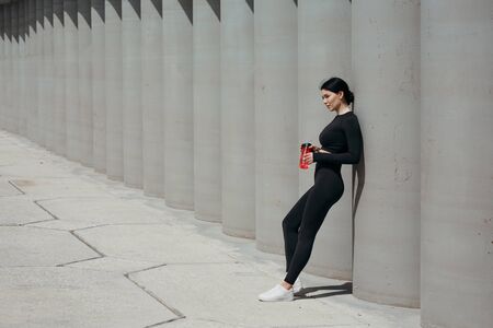 Horizontal photo of an Asian girl playing sports on the street and leaning on a wall holding a water...