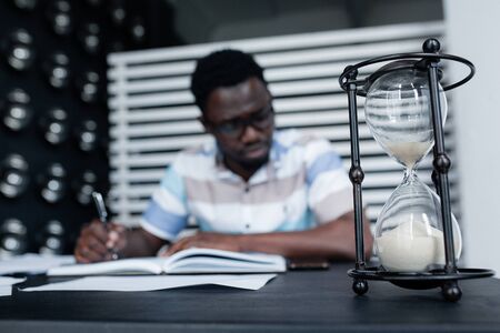 A Ethiopian black student works hard at textbooks before the exam week begins. Hourglass on the background.の写真素材
