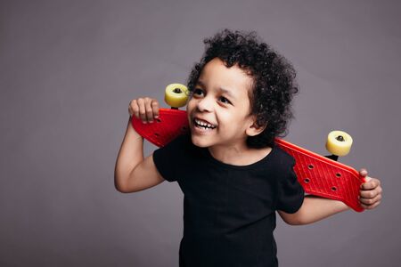 Close-up of a curly dark skinned boy in a black T-shirt holding red skateboard behind his back and smiling on gray backgroundの写真素材