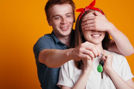 Young man closes his eyes to the girl with his palm and holds keys to new car in front of her. Guy...