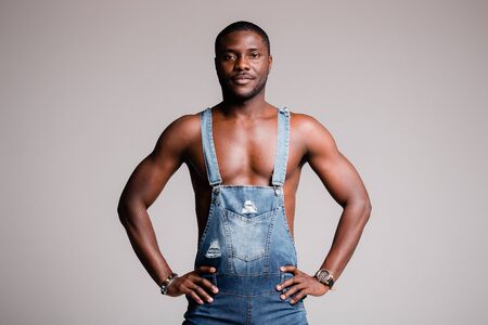 Portrait of a dark-skinned Frenchman with a naked torso wearing a denim jumpsuit and rests his hands...