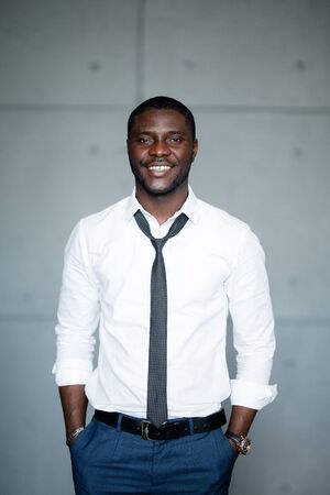 Portrait of a black man in a white shirt standing with his hands in his pockets and smiling at the...