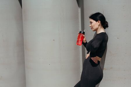 Rear view of a girl athlete who put her hand on her side and drinks water from a red drinking bottle while resting after a workoutの写真素材