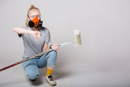 Girl painter with a paint roller construction in orange hands and a respirator on her face. Girl is dissatisfied with paint.の写真素材