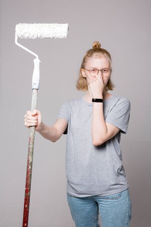 Blonde girl holding a paint roller in his hand and pinches his nose. Attractive girl complain about the smell of paint.の写真素材