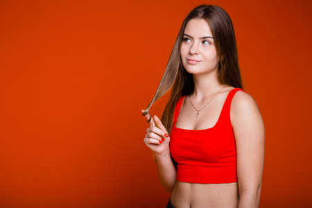 An American in a red topic looks up thoughtfully and winds a lock of long hair over her finger on an orange backgroundの写真素材