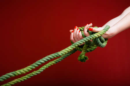 Female hands are tangled in a green rope on a burgundy background. The concept of problems that pull down and the inability to get rid of themの写真素材