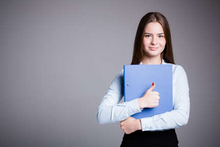 Girl office worker holds a blue folder in her hands and shows thumb up in approval and like on a gray backgroundの写真素材