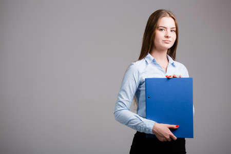 Business portrait of a young woman in a blue shirt and black skirt who holds a folder with documents in her hands on a light backgroundの写真素材