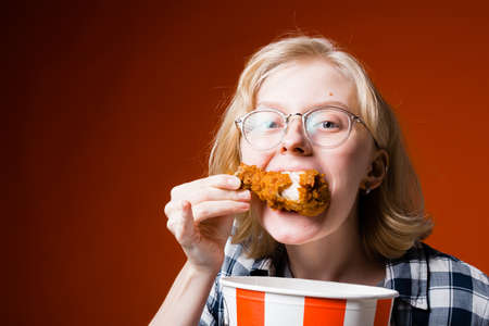 Girl blonde in glasses with an appetite eats a big chicken leg on a red background. Fast food bannerの写真素材