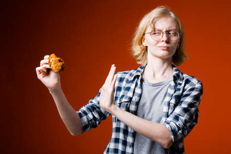 A girl of European appearance holds a chicken leg in her hand and holds her hand in front of her with a displeased face as a sign of rejection of harmful food on a red backgroundの写真素材