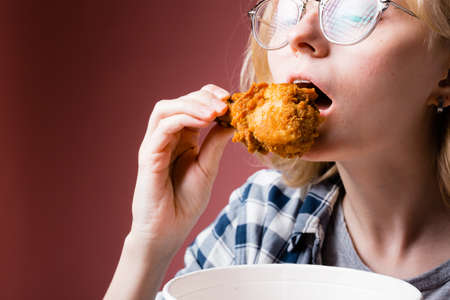 Closeup portrait of a blonde girl who brings a chicken leg to her mouth while holding a bucket of chicken in her handsの写真素材