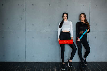 Two beautiful female athletes are standing against the background of a gray wall with Pilates equipment. Fitness trainer holding massage roll and training rubber bandの写真素材