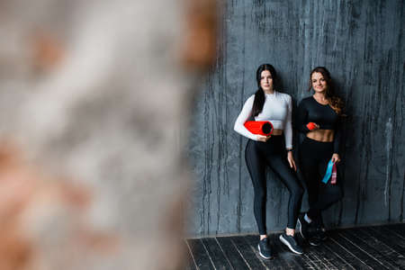 Two beautiful female athletes are standing against the background of a gray wall with Pilates equipment. Fitness trainer holding massage roll and training rubber bandの写真素材