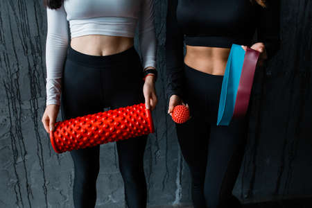 Close up. Two female are standing against the background of a gray wall with Pilates equipment. Fitness trainer holding massage roll, massage ball and training rubber bandの写真素材
