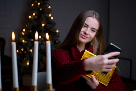 Attractive Caucasian girl sitting at a festive Christmas table, hugging a gift wrapped box and...