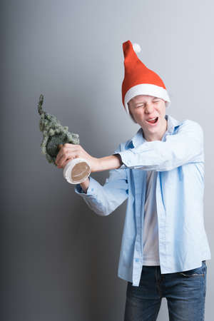 A young guy in a red Christmas hat and blue shirt stands with a miniature of a Christmas tree on a gray background. Teenager having fun and getting ready for christmasの写真素材