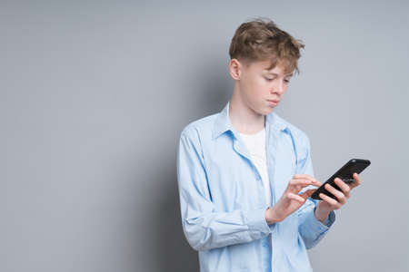 A teenager boy in a blue shirt and a white T-shirt plays the phone on a gray background. Troubled teenager, social issues, gamer, phone addictionの写真素材