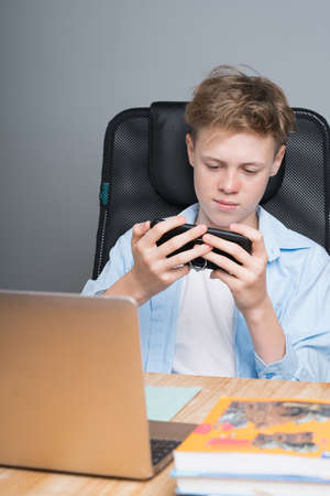 Caucasian boy sits at a desk, plays on the phone, sitting on an online lesson. Homeschooling,...