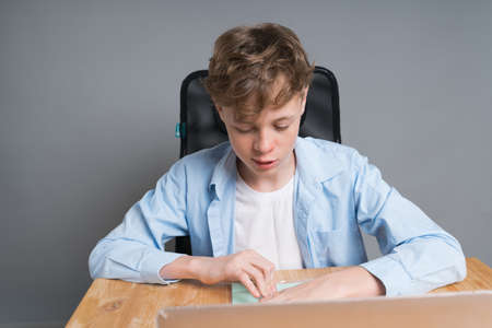 A young boy student in a blue shirt sits at an online lesson, at a table waiting for the start of...