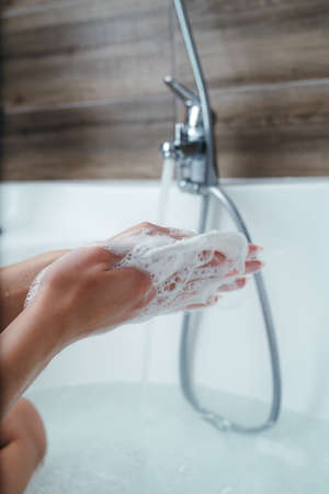 The girl is foaming a washcloth under running water from the tap, sitting in the bath. close-up of...