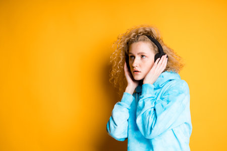 A girl with blond hair looks scared to the side, pressing headphones to her ears