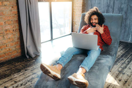 An african american man in a red plaid shirt talking by video call with friends on a laptop, sits in...