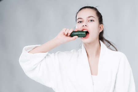 A beautiful young girl in a white robe brings a cucumber to her mouth. Healthy eatingの写真素材