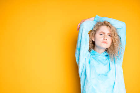 A young girl in a blue sweater raises her hands up behind her head on a bright backgroundの写真素材
