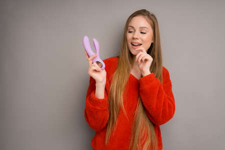A dildo in the hands of an attractive woman in a red sweater who makes a flirtatious expression. Isolated on a gray background. Gift concept for March 8の写真素材