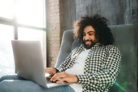 Smiling Egyptian in a plaid shirt sits in a chair working at the computer. Work remotelyの写真素材