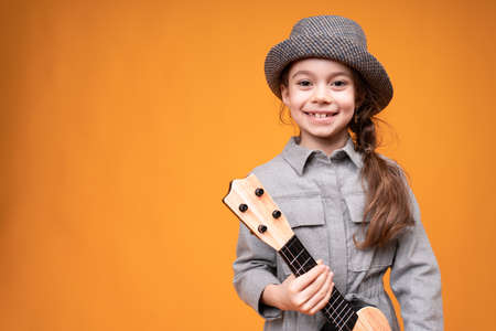 Adorable child 8-9 years old in a hat stands on a bright background holding a ukulele in his handsの写真素材