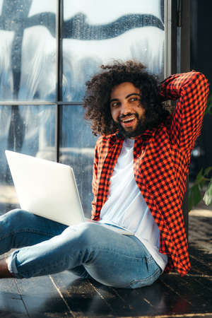 Handsome african american man in red plaid shirt sitting at laptop with one hand behind his headの写真素材