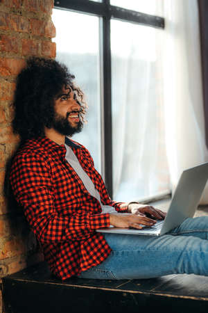 African American man in jeans sits on the floor against the background of a window at a computer