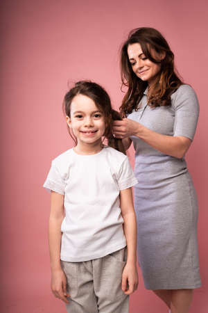 Beautiful woman in a long dress braids her little daughters hair while standing on a pink backgroundの写真素材