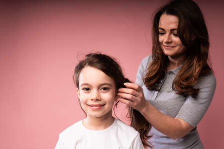 Beautiful woman in a long dress braids her little daughters hair while standing on a pink background