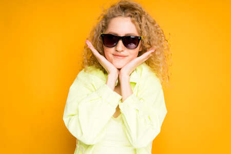 Cute blonde girl with curly hair wearing glasses leans her hands to her face smilingの写真素材