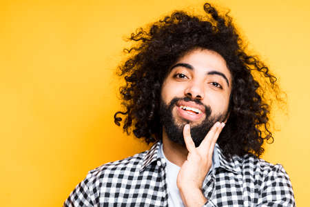 Cute young african american man with black beard smiles and touches his hand to his faceの写真素材