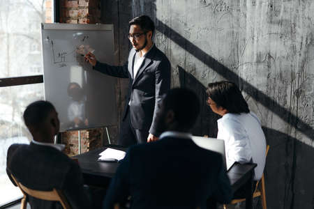 A designer in a suit stands near a whiteboard with drawings talking to other workers at the table. Cabinet meetingの写真素材