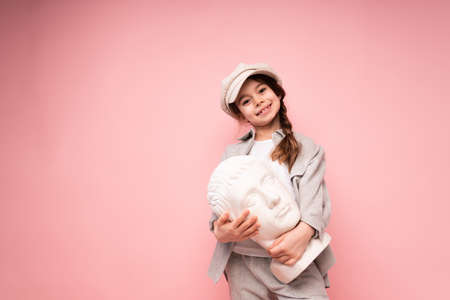 Cute little girl with a pigtail holding a sculpture in the shape of a head standing on a pink backgroundの写真素材