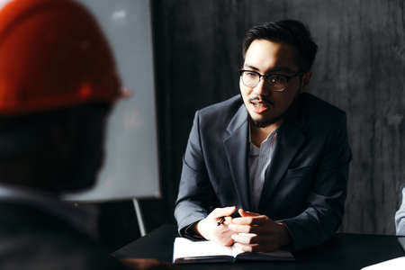Korean worker in suit interviewing African American worker in construction helmet sitting next to drawing boardの写真素材