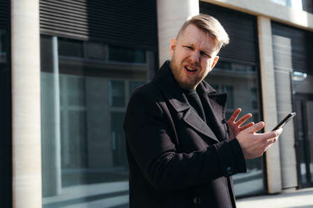 Stylish businessman in a black coat holds a phone in his hands and is perplexed and disappointed...