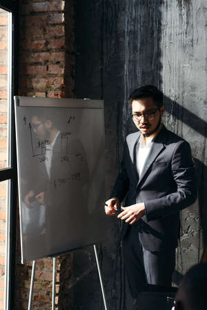 Vietnamese man with glasses stands near a white board with a schedule holding a conference in the officeの写真素材