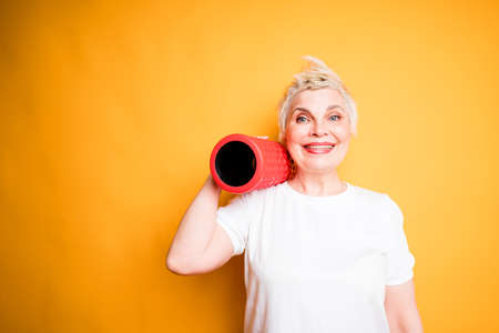 an elderly woman holds a fascia roll on her shoulder and rejoices after a fitness workout. yellow background for textの写真素材