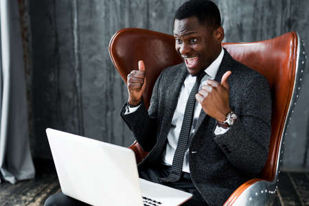 an investor from nigeria rejoices at the profit and shows a thumb up and looks at a laptop while sitting in a chair. finance conceptの写真素材