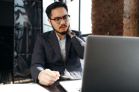 Tired asian businessman in a suit sits at a table and works at a laptop while doing a neck massage with his hand because of fatigueの写真素材