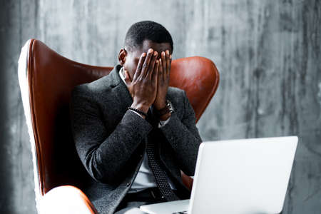 Tired african american office worker sits on a chair at a laptop and covers his face with his hands