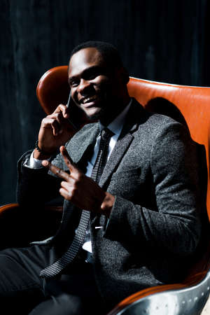 Handsome african american businessman sitting on chair and talking on the phone showing peace signの写真素材