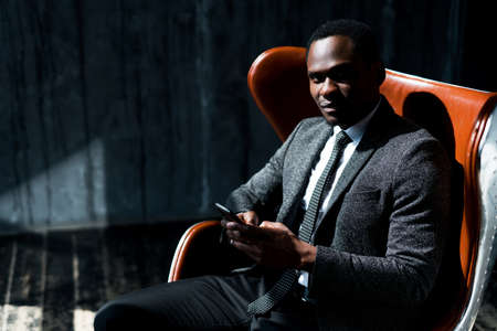 Smiling african american businessman sitting on chair and working on phone while looking at cameraの写真素材